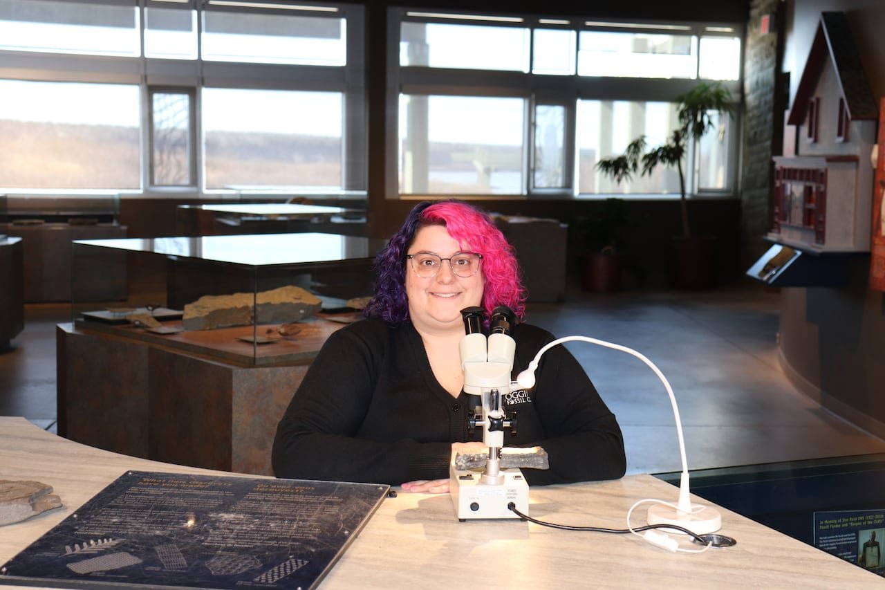 A woman in a black top sits behind a microscope in a large exhibit space.