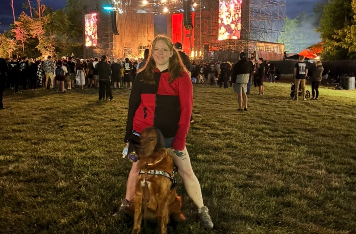 A woman stands in the crowd at an outdoor concert, her golden retriever standing between her legs.